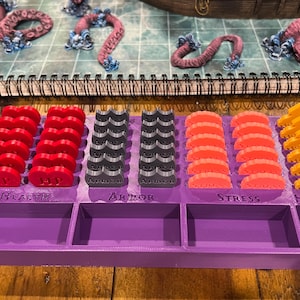 May include: A purple game organizer with labeled compartments for Health, Armor, Stress, and Hope. It holds red, gray, orange, and yellow game pieces. The game pieces are designed to look like health points, armor, stress, and hope.
