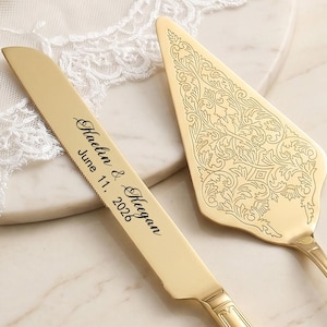 May include: A gold-colored cake knife and server set. The knife is engraved with the names "Kaelin & Keegan" and the date "June 11, 2026." The server has an ornate floral design. The set is displayed on a marble surface.