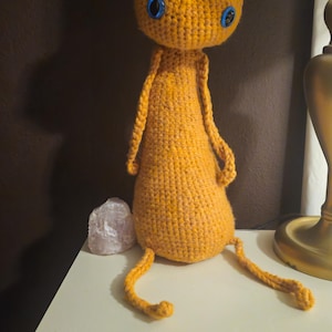 May include: A handmade, orange crochet doll with large blue eyes. The doll has a rounded head, elongated body, and dangling arms and legs. It is sitting on a white surface, with a crystal and lamp in the background.
