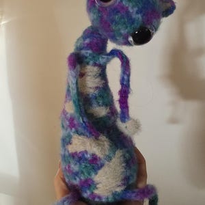 May include: A handmade plush creature crafted from variegated yarn in shades of purple, teal, and blue with patches of cream-colored faux fur. The figure features two large, glossy black button eyes, a long neck, and thin, dangling limbs.