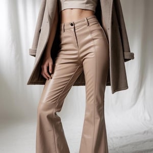 May include: Tan faux leather trousers with a flared leg, worn with a matching crop top and a beige coat. The trousers have a high waist and visible stitching details. The outfit is a neutral colour.