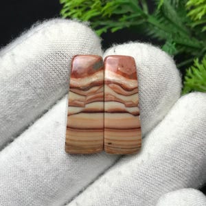 Op de afbeelding: Twee rechthoekige, gepolijste edelstenen met een gelaagd patroon van bruin, beige en wit. De stenen worden tegen een witte stoffen achtergrond gehouden, die hun natuurlijke kleuren en texturen laat zien. De edelstenen hebben een gladde, glanzende afwerking.