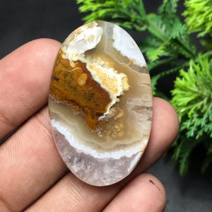 Puede incluir: Gema ovalada con un patrón natural de capas marrones, blancas y beige. La piedra pulida presenta una superficie lisa con áreas opacas y translúcidas. La piedra se sostiene en una mano, mostrando sus tonos terrosos y su diseño único.