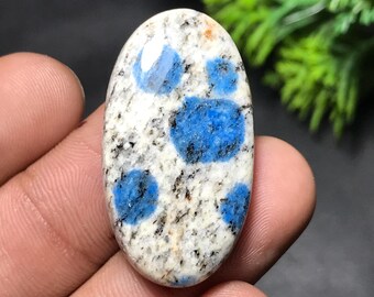 May include: Oval-shaped K2 stone with a white base, speckled with black and featuring vibrant blue spots. The polished gemstone is held in a hand, showcasing its natural patterns and colors. The stone's smooth surface reflects light, highlighting its unique mineral composition.
