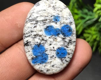 May include: Oval-shaped K2 stone, also known as K2 jasper, featuring a white base with black speckles and vibrant blue orb-like patterns. The polished gemstone is held in a hand, showcasing its natural beauty.