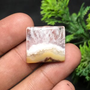 Op de afbeelding: Een vierkante edelsteen met een gelaagd uiterlijk. De bovenste laag is doorschijnend met witte slierten, gevolgd door een gele band en een bruine basis. De steen wordt in een hand gehouden.