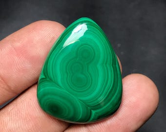 May include: A polished, teardrop-shaped malachite gemstone. The vibrant green stone features concentric, swirling patterns, creating a unique and natural design. The gemstone is held in a hand, showcasing its smooth, reflective surface.