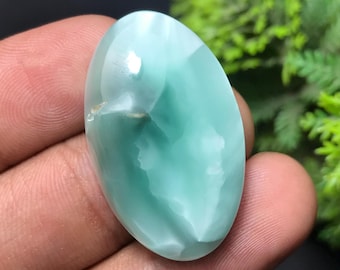 May include: A polished, oval-shaped gemstone with a light teal color and a glossy finish. The stone has a swirling pattern of white and teal, with a small, natural inclusion. The gemstone is held in a hand, with green foliage in the background.