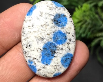 May include: Oval-shaped K2 stone cabochon. The stone features a white base with speckled black inclusions and vibrant blue spots. The polished surface highlights the natural patterns and colors of the gemstone.