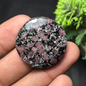 Könnte beinhalten: Ein runder, polierter Edelstein mit einem gesprenkelten Muster. Der Stein weist eine Mischung aus schwarzen, weißen und rosa Farbtönen auf, wobei die rosa Elemente als kleine, kreisförmige Einschlüsse erscheinen. Die Oberfläche ist glatt und reflektierend.