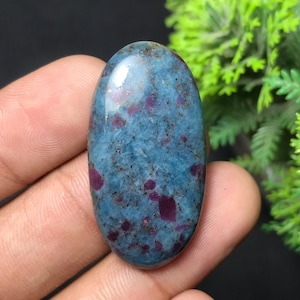 Peut inclure: Pierre précieuse de forme ovale avec une surface polie. La pierre présente une couleur de base bleue avec des inclusions rouges dispersées. De petits points bruns sont visibles. La pierre est tenue dans une main.