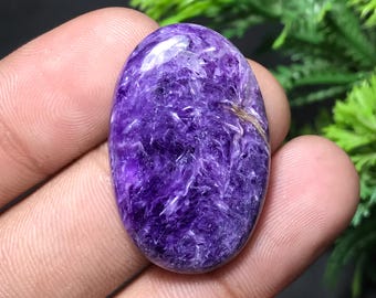 May include: Oval-shaped polished gemstone, predominantly purple with white and gold veining. The stone is held in a hand, showcasing its smooth, reflective surface. The gemstone's color ranges from deep violet to lighter shades.