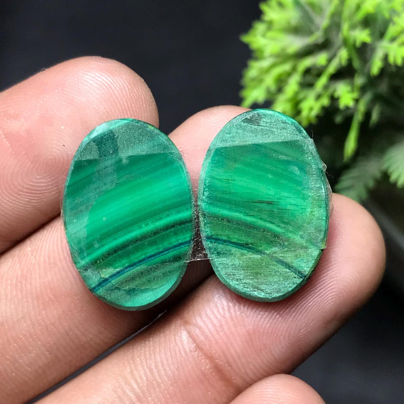 May include: Two oval-shaped malachite gemstones. The gemstones are a vibrant green with darker green and blue-green banding. The stones are polished and have a smooth surface. The background is dark, with a hint of green foliage.