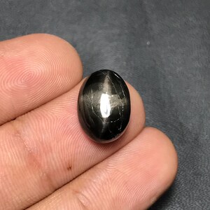 Puede incluir: Una gema negra pulida, de forma ovalada, con un efecto de estrella. La piedra tiene una superficie brillante con una prominente línea blanca reflectante en su centro. La gema es de color oscuro y opaco.