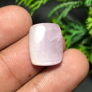 May include: A polished, rectangular gemstone with rounded edges. The stone has a soft, pale pink hue with a subtle, translucent quality. The gemstone is held in a hand, with a blurred green background.