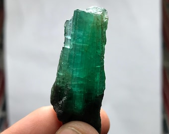 Puede incluir: Un cristal de esmeralda en bruto y alargado que presenta un tono verde vibrante y translúcido con estrías verticales visibles y facetas naturales. La base del espécimen mineral es más oscura y opaca, hacia una sección superior más clara.