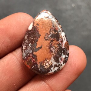 May include: A teardrop-shaped gemstone with a mix of colors. The stone features a prominent area of copper-orange, surrounded by dark brown, white, and hints of red. The polished surface reflects light, highlighting the natural patterns.