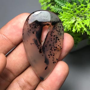 May include: A teardrop-shaped gemstone with a polished surface. The stone is a mix of translucent white and brown hues, speckled with black inclusions. A natural cavity runs through the center. The gemstone is held in a hand.