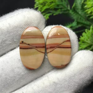 以下が含まれることがあります： 磨かれた楕円形の宝石が2つ。ベージュ、オレンジ、茶色の帯が層状に重なった模様です。各石の表面には細い暗い線があります。宝石は白い背景に置かれています。