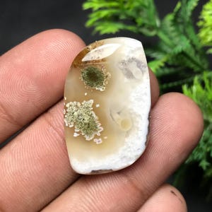 May include: A polished gemstone with a creamy white base, featuring areas of tan and green inclusions. The stone has a rounded, irregular shape and a smooth, glossy surface. The gemstone is held in a hand.