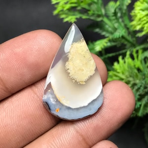 Puede incluir: Una gema pulida en forma de lágrima. La piedra presenta capas de blanco, gris y azul claro, con un parche central de color amarillo-beige. La gema se sostiene en una mano, con follaje verde en el fondo.