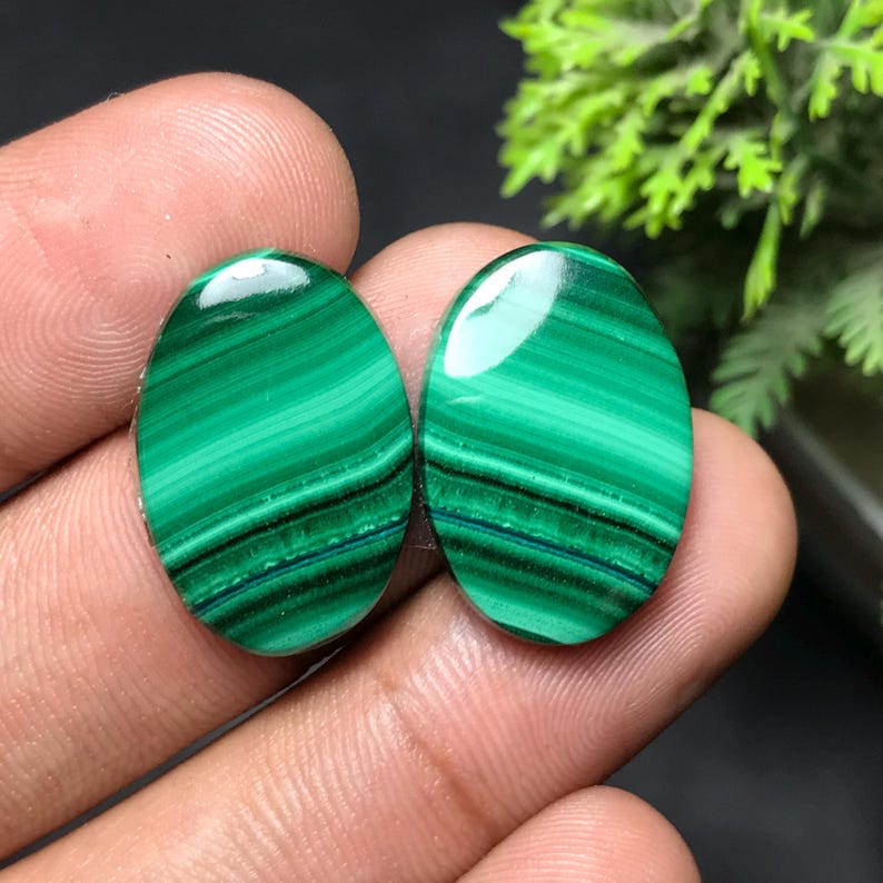May include: Two polished oval malachite gemstones. The stones display a vibrant green color with dark green and black banding patterns. The gemstones are held in a person's hand, with a blurred background.