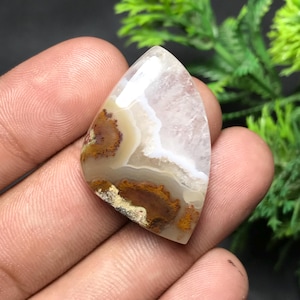 Puede incluir: Una piedra preciosa pulida con un patrón único. La piedra presenta una mezcla de colores, incluyendo blanco, beige y marrón, con intrincadas vetas y patrones. La forma es un triángulo redondeado, mostrando la belleza natural de la piedra.