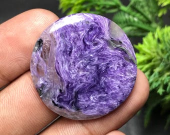 May include: A round, polished charoite gemstone. The stone features swirling patterns of deep purple, lavender, and translucent areas, with hints of black and green. The gemstone is held in a hand, with green foliage in the background.