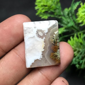 May include: A rectangular gemstone with a unique pattern. The stone features a blend of white, brown, and green hues, with a marbled appearance. The colors are swirled together, creating an organic, natural design. The stone is held in a hand.