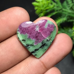 Pode incluir: Pedra preciosa em forma de coração com uma paleta de cores vibrantes. A pedra apresenta um centro magenta profundo, rodeado por uma faixa verde com inclusões pretas. A superfície polida reflete a luz, realçando os padrões naturais.