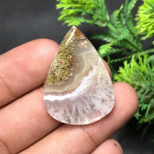 May include: A teardrop-shaped gemstone with a distinctive pattern. The top section displays brown and green shades, fading into white and lavender towards the base. The stone has a polished, translucent look, revealing natural mineral formations.