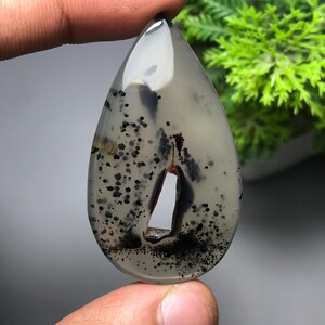 May include: A teardrop-shaped gemstone with a polished surface. The stone is primarily gray with black speckles and inclusions. A large, irregular opening is visible near the bottom. The gemstone is held between two fingers.