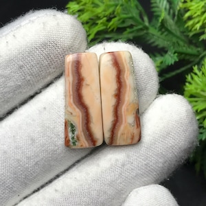 Op de afbeelding: Twee rechthoekige edelstenen met een gepolijste afwerking. De stenen hebben een crèmekleurige basis met wervelende bruine en oranje patronen. Enkele groene en witte insluitsels zijn zichtbaar. De stenen worden op een wit oppervlak gepresenteerd.