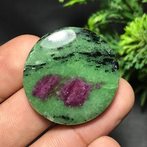 May include: A circular, polished gemstone with a vivid green hue, black streaks, and two striking magenta patches. The stone is held in a hand, highlighting its natural beauty and unique pattern.