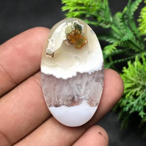 Può includere: Pietra preziosa ovale con un aspetto a strati. La sezione superiore presenta una base bianco crema traslucida con macchie verdi, arancioni e marroni. Sotto, uno strato bianco a forma di nuvola si trova sopra una sezione di viola chiaro e bianco. Il fondo è bianco pieno.