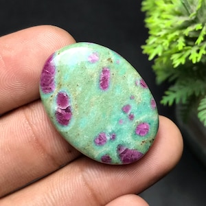 May include: An oval gemstone featuring a light green base with magenta inclusions. The polished stone has a smooth surface and a natural appearance. The gemstone is held in a hand, with a blurred green plant in the background.