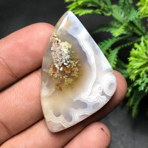 May include: A polished, triangular gemstone with a unique pattern. The stone features swirling white, gray, and tan bands, with a central area of mossy green and reddish-brown inclusions. The overall effect is a natural, organic aesthetic.