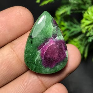 Op de afbeelding: Een gepolijste, druppelvormige edelsteen. De steen vertoont een opvallende combinatie van kleuren, met een diepgroene basis en een levendig magenta centrum. Het patroon is een mix van de twee kleuren, wat een unieke visuele textuur creëert.