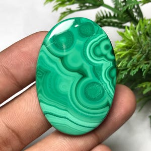 May include: Oval-shaped polished malachite gemstone. The stone displays a vibrant green color with swirling patterns and concentric circles. The polished surface reflects light, highlighting the natural variations in the stone's texture.