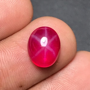 Op de afbeelding: Een ovale ster-robijn edelsteen. De dieprode edelsteen heeft een zesstralig sterpatroon. De ster is helderwit tegen de rode achtergrond. De steen is gepolijst.
