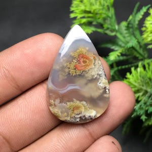 Pode incluir: Uma pedra preciosa em forma de gota com um padrão branco, cinza e marrom. A pedra apresenta uma ponta branca e padrões internos intrincados com toques de laranja e amarelo, assemelhando-se a uma paisagem ou design abstrato.