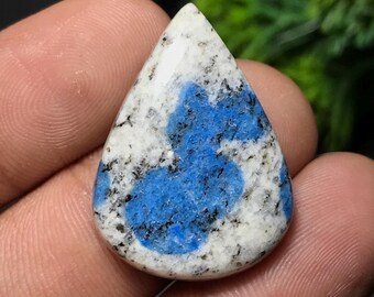 May include: A polished teardrop-shaped gemstone with a white base, speckled with black and featuring a vibrant blue pattern in the center. The stone is smooth and reflective, showcasing natural mineral patterns.