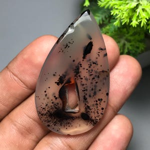 May include: A teardrop-shaped gemstone pendant with a translucent, light gray base. The stone features black speckles and inclusions, with a hint of orange near the center. The pendant is held in a hand.