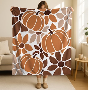 May include: A large, rectangular blanket featuring a repeating pattern of stylized pumpkins and flowers in shades of brown, orange, and white. The blanket is held up, showcasing the full design and color palette.