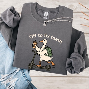 May include: Charcoal grey sweatshirt featuring the phrase "Off to fix teeth" and a cartoon goose on a scooter, holding a coffee cup. The goose has a backpack. A silver necklace is draped on the sweatshirt.