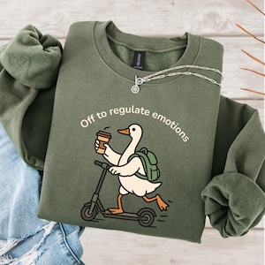 May include: An olive green sweatshirt with the phrase "Off to regulate emotions" printed above a cartoon duck riding a scooter and holding a coffee cup. The duck has a backpack. A silver necklace is draped across the sweatshirt.