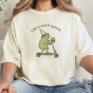 以下が含まれることがあります： クリーム色のTシャツ。魔法使いの帽子をかぶり、コーヒーカップを持ち、スクーターに乗っている緑色のカエルの気まぐれなイラスト。テキスト「Off to teach speech」はカエルの上にアーチ状に配置されています。