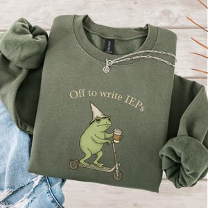 Könnte beinhalten: Olivgrünes Sweatshirt mit dem Text "Off to write IEPs" und einem Cartoon-Frosch mit Zaubererhut und Sonnenbrille, der einen Roller fährt und einen Kaffeebecher hält. Eine silberne Halskette ist ebenfalls zu sehen.