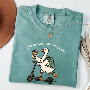 May include: Teal Comfort Colors t-shirt with a graphic of a duck riding a scooter and holding a coffee cup. The text "Off to overthink everything" is printed above the duck. A silver necklace with a pendant is also visible.