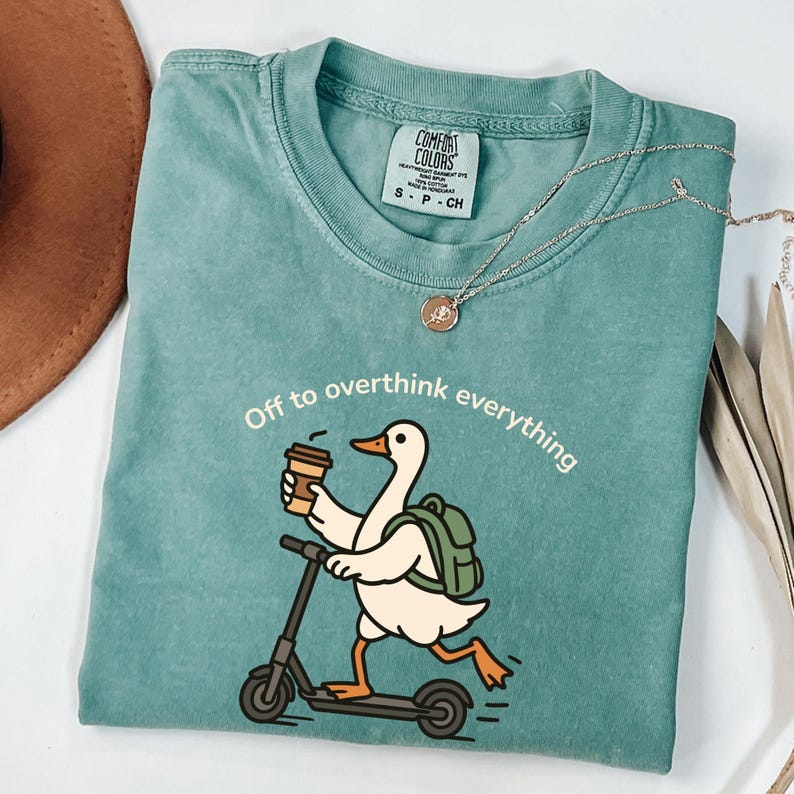 May include: Teal Comfort Colors t-shirt with a graphic of a duck riding a scooter and holding a coffee cup. The text "Off to overthink everything" is printed above the duck. A silver necklace with a pendant is also visible.
